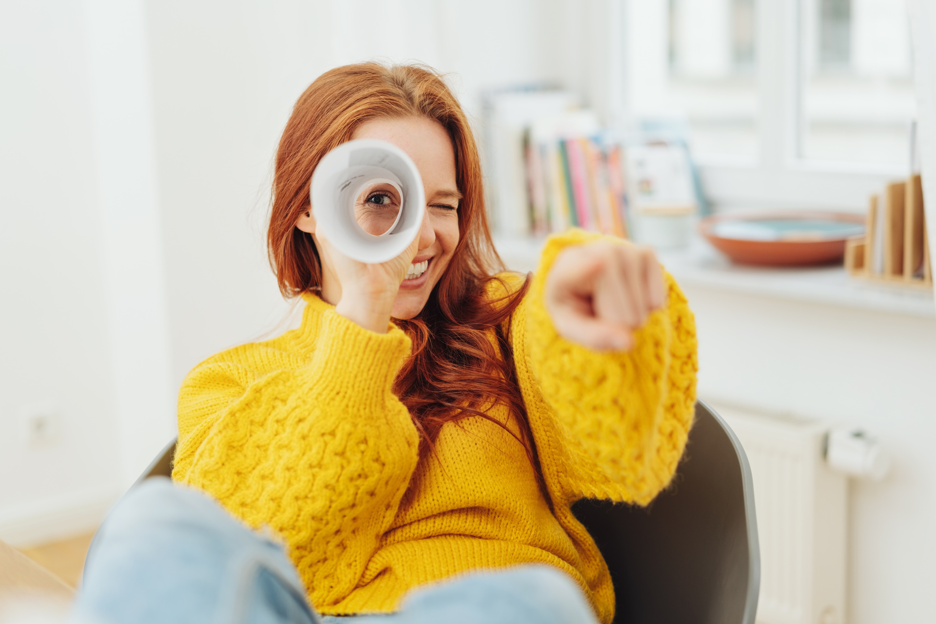 Eine junge rothaarige Frau im gelben Pulli hält ein zusammengerolltes Blatt Papier vor ein Auge wie ein Fernrohr und zeigt mit dem Finger auf den Betrachter.