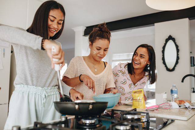 Drei junge Frauen stehen nebeneinander in der Küche und kochen gemeinsam – sie lachen und haben sichtlich Spaß.