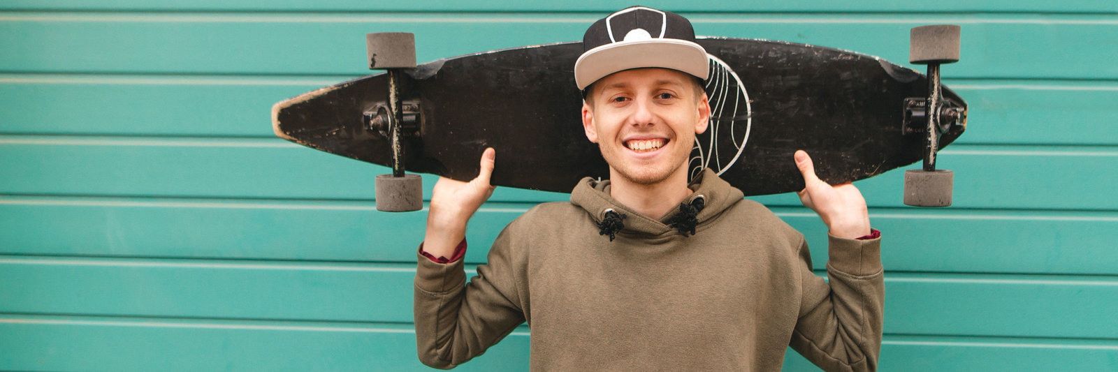Ein junger Mann mit Hoodie und Cappy steht vor einer grünen Wand und trägt ein Skateboard lässig auf den Schultern.