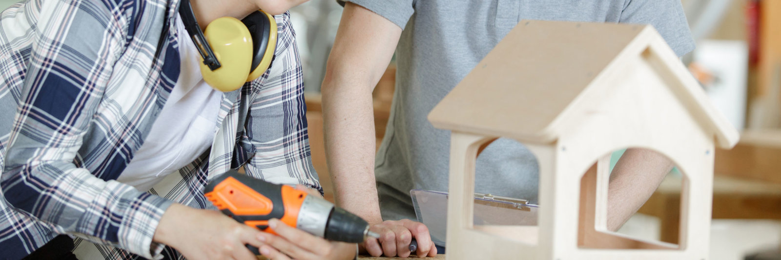 Zwei Personen bauen gemeinsam ein kleines Holzhäuschen – im Bild sind nur ihre Hände und ein Teil des Körpers zu sehen.