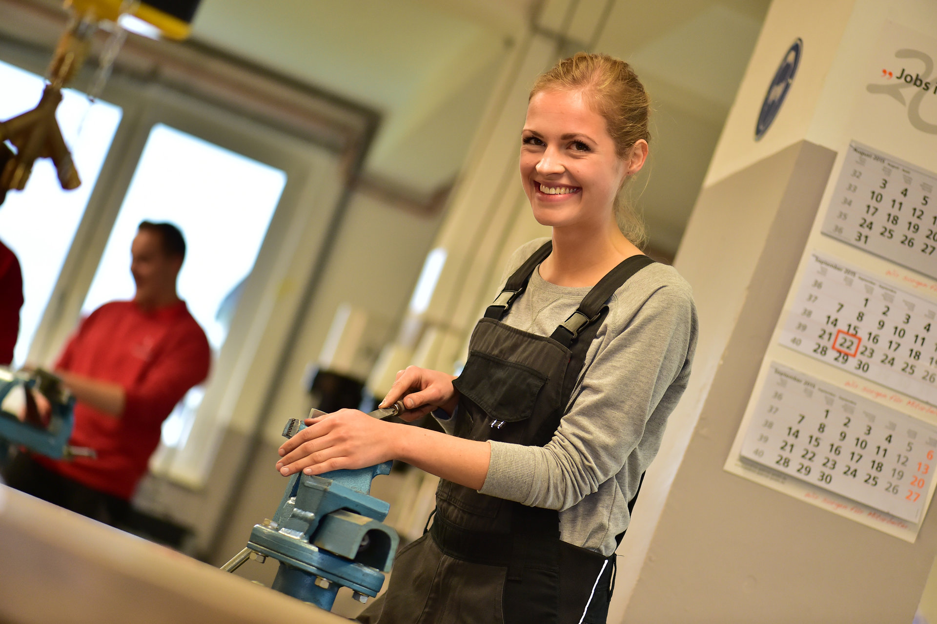 Eine Teilnehmerin in der Werkstatt hält eine Feile in der Hand und lächelt in die Kamera, während ein Werkstück im Schraubstock eingespannt ist.