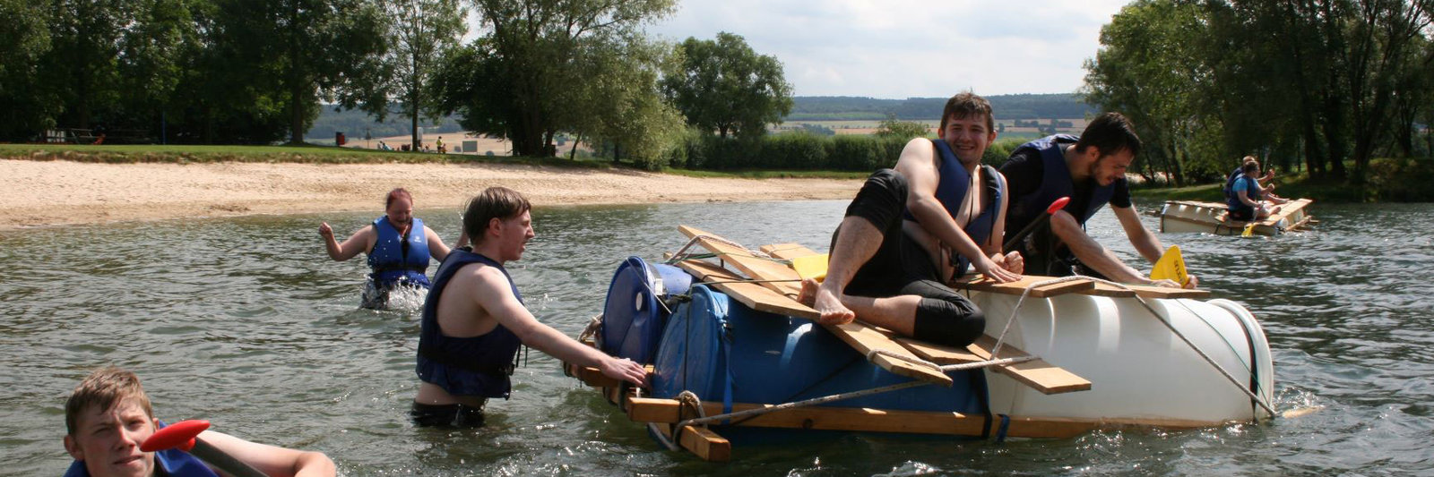 Zwei Teilnehmende sitzen auf einem Floßboot auf dem See, eine Person steht im Wasser und hält das Floß fest – alle tragen Schwimmwesten.