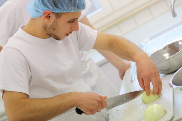 Ein Teilnehmer der Küche trägt eine Kopfhaube und schneidet sorgfältig eine Zwiebel.