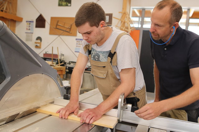 Ein Teilnehmer arbeitet an der Tischkreissäge und sägt ein Stück Holz. Der Ausbilder steht dahinter und beobachtet den Arbeitsvorgang aufmerksam.