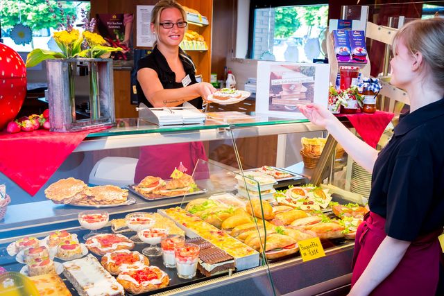 Zwei Teilnehmerinnen stehen an einer gut gefüllten Auslage im Café – eine hinter der Theke, die andere davor.