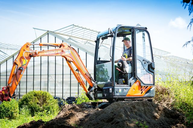 Ein Teilnehmer bedient bei sonnigem Wetter einen kleinen Bagger auf dem Gelände des KBBW. Im Hintergrund ist ein Gewächshaus zu sehen.
